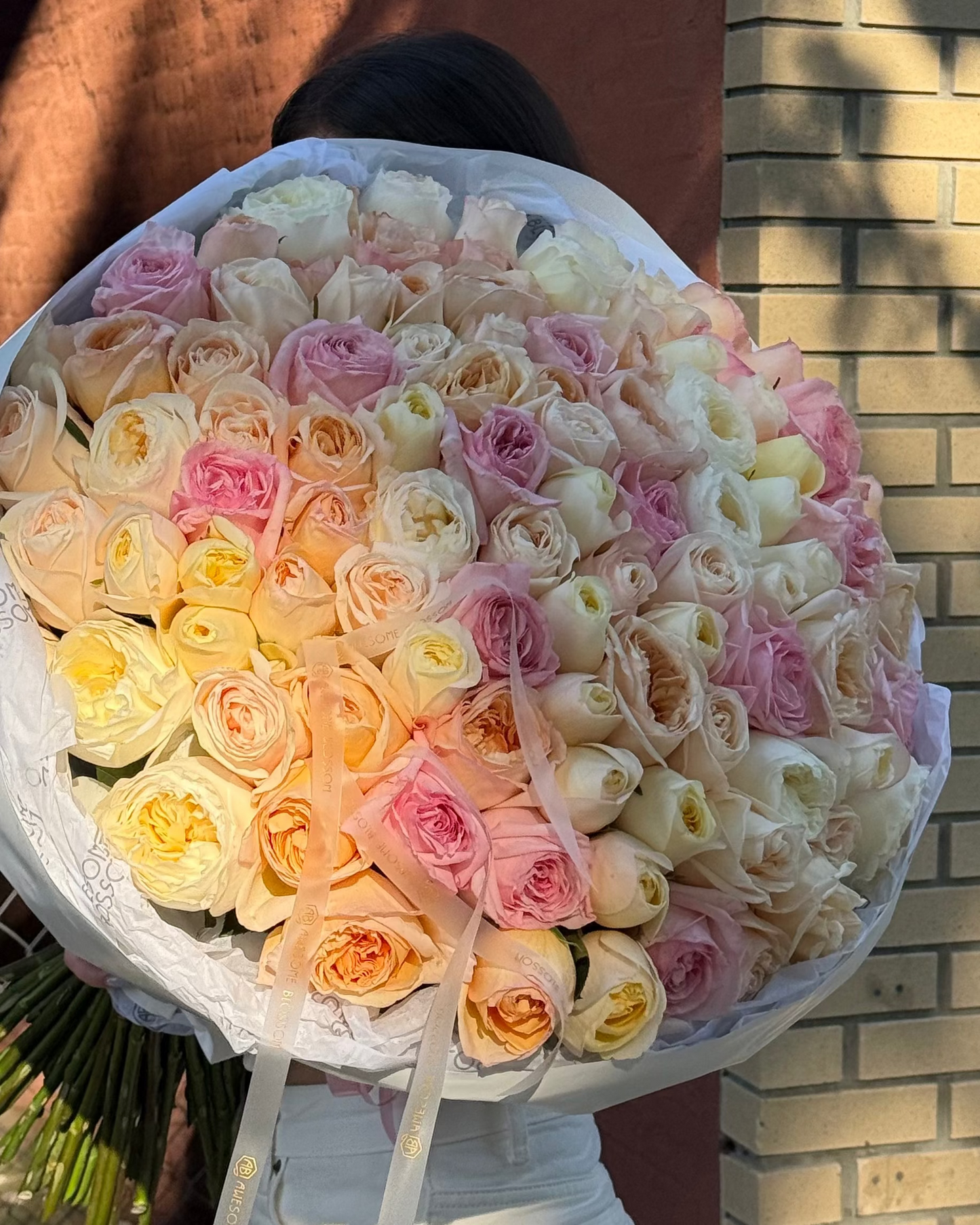 A large, round bouquet of multicolored roses in shades of pink, yellow, orange, and cream, held by a person wearing white pants in front of a brick wall.