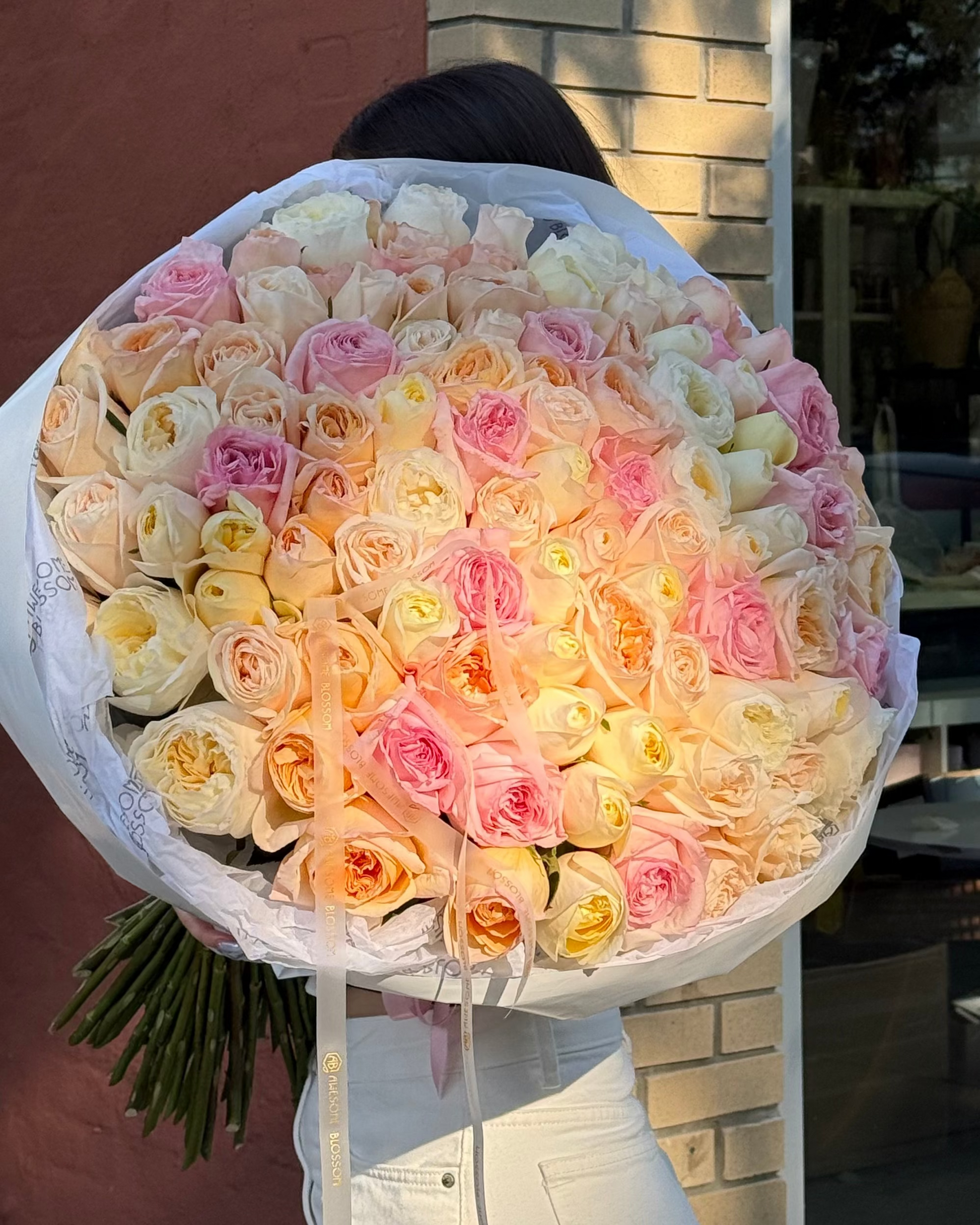 A large bouquet of fresh roses wrapped in white paper, held by a person standing in front of a storefront with a brick column and glass window.