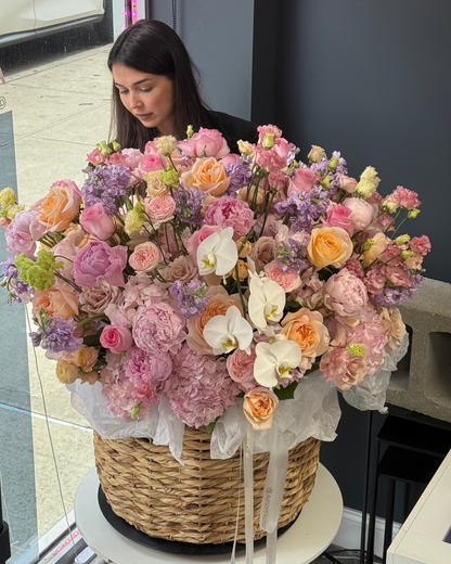 A large, vibrant pastel flower arrangement in a woven wicker basket, displayed on a small white table inside a shop. A woman is visible in the background near the window.