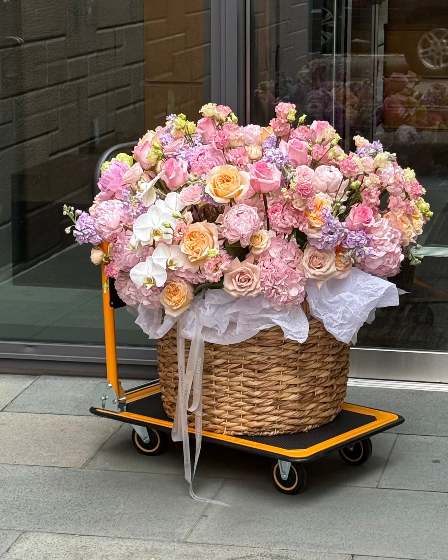 A large floral arrangement in a woven basket on a yellow and black dolly cart, parked outdoors on a stone sidewalk in front of a glass storefront.