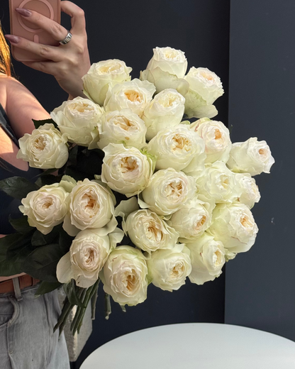 A large bouquet of cream-colored roses held by a person taking a mirror selfie in a room with a dark gray wall and a white round table.