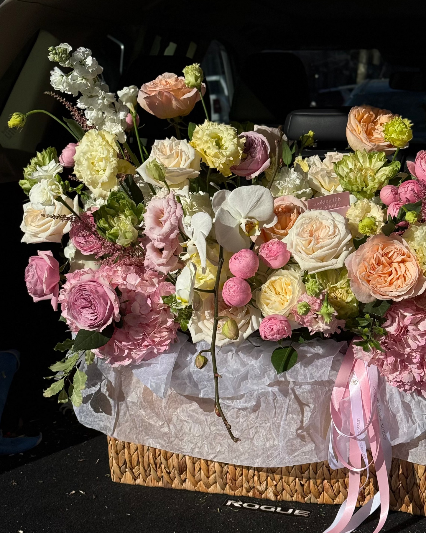 Floral arrangement in a woven basket with pink ribbon on a car trunk.